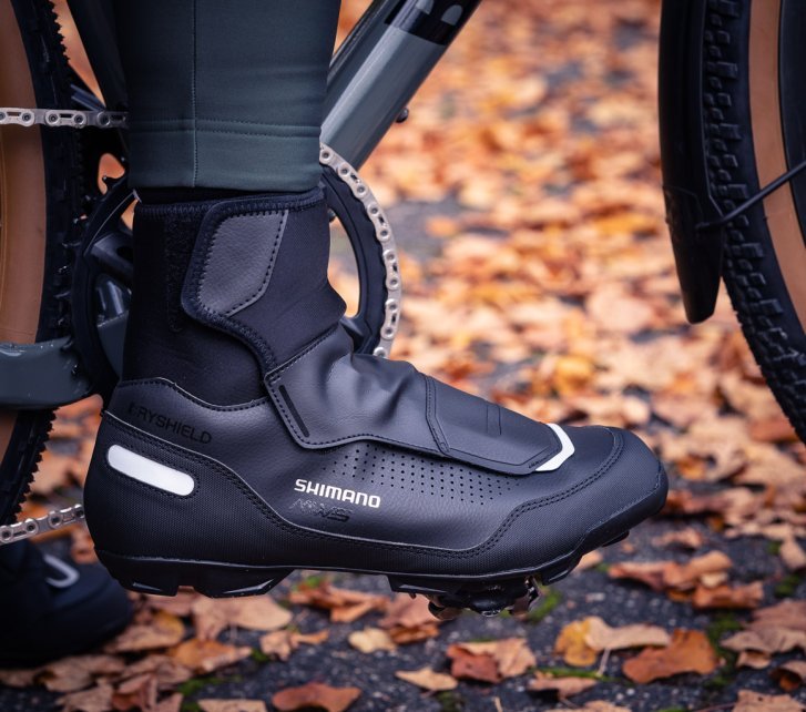 Pie con zapatilla ciclista invernal negra sobre un pedal, hojas otoñales al fondo.