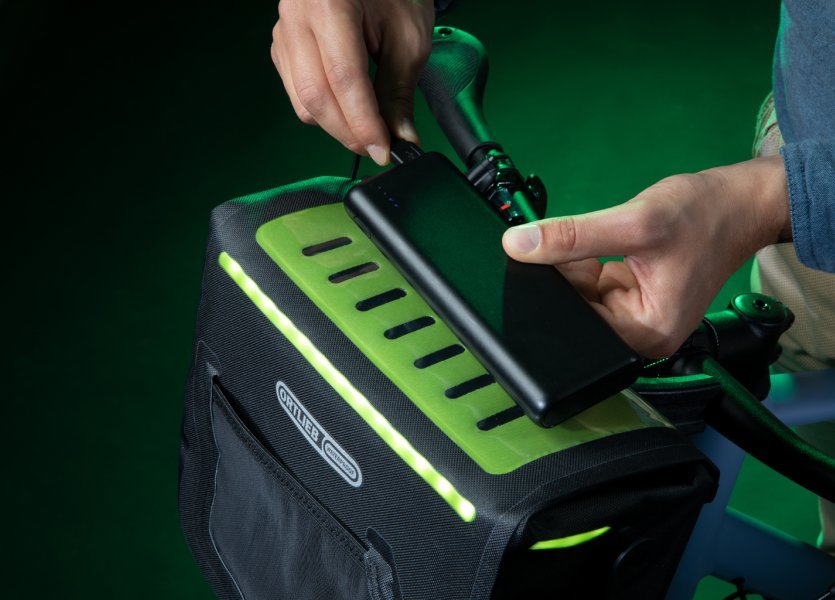 Zwei Hände: Eine hält ein Smartphone, das an eine schwarze Fahrradtasche mit grünen Akzenten angeschlossen ist, die andere berührt es. Die Tasche ist am Lenker befestigt und hat ein beleuchtetes, perforiertes Design. Der Hintergrund ist dunkelgrün.