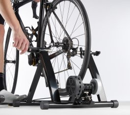 A hand secures a road bike onto a black indoor trainer with a roller.