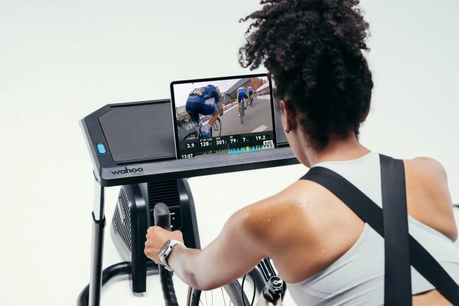 Primer plano: over-shoulder – Una mujer de pelo oscuro entrena en un rodillo en un estudio fotográfico blanco. Frente a ella hay una tablet en la que se muestra la plataforma de entrenamiento Wahoo SYSTM.