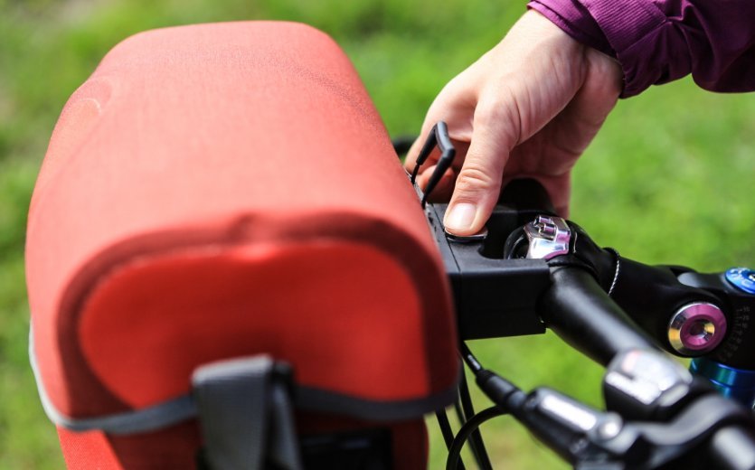 A hand is opening or closing a red Ortlieb bicycle bag attached to a bike. Part of the handlebar is visible, with a blurry green field in the background.