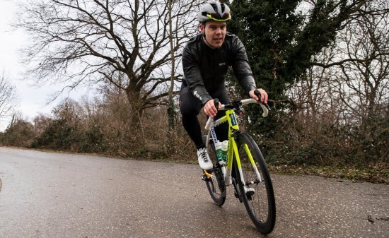 A cyclist on a black and yellow road bike rides on a wet road, surrounded by bare trees and bushes on a cloudy day.