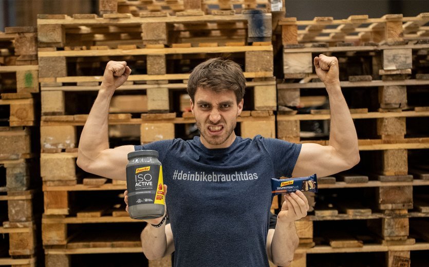 A man flexes his muscles. Behind him, another man is holding various nutritional supplements up to the camera.