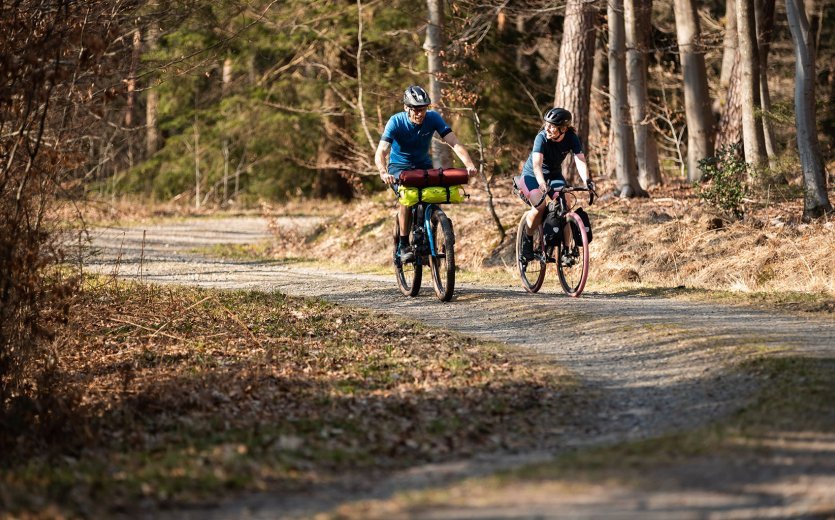 Rainer und Svenja von bc fahren mit ihren bepackten Bikes über einen Waldweg. Es ist sonnig.