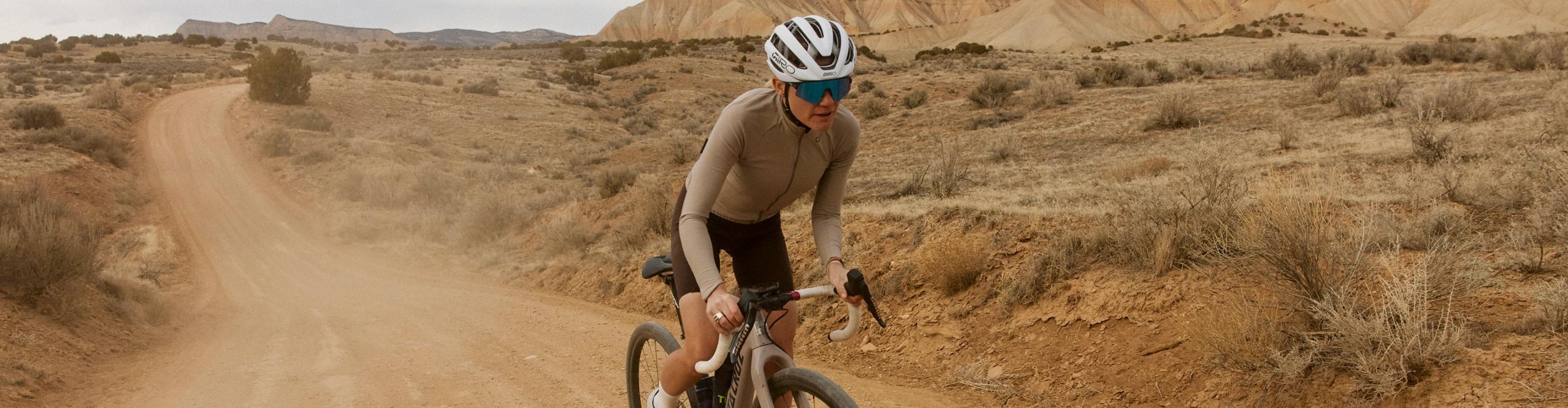 Gravel-Fahrerin mit weißem Giro Fahrradhelm fährt auf staubiger Schotterstraße durch eine weite Wüstenlandschaft.