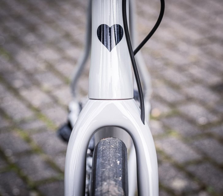 Detailfoto: Ein breiter Straßenreifen benötigt viel Platz in der Gabel eines hellgrauen Blackheart Rennrads. Das Steuerrohr ziert ein schwarzes Herz, im Hintergrund eine grau plattierte Fläche 