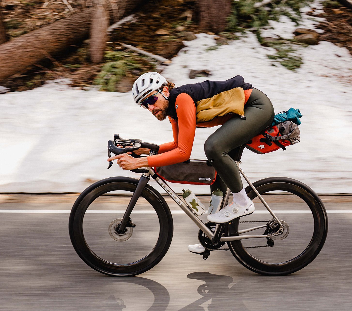 Rennradfahrer in Assos-Bekleidung fährt dynamisch durch verschneite Berglandschaft mit voll bepacktem Bikepacking-Setup.