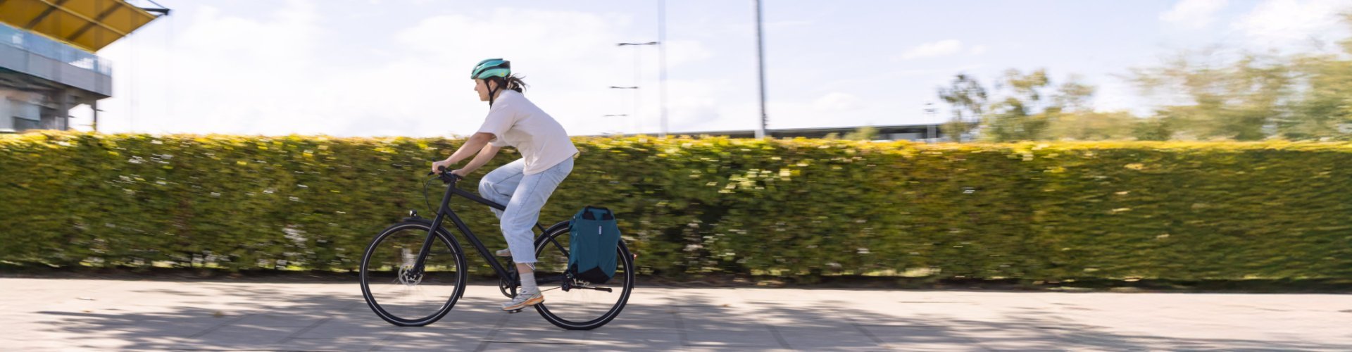 A person rides a bicycle with a helmet along a green hedge, with a large bag on the luggage rack.