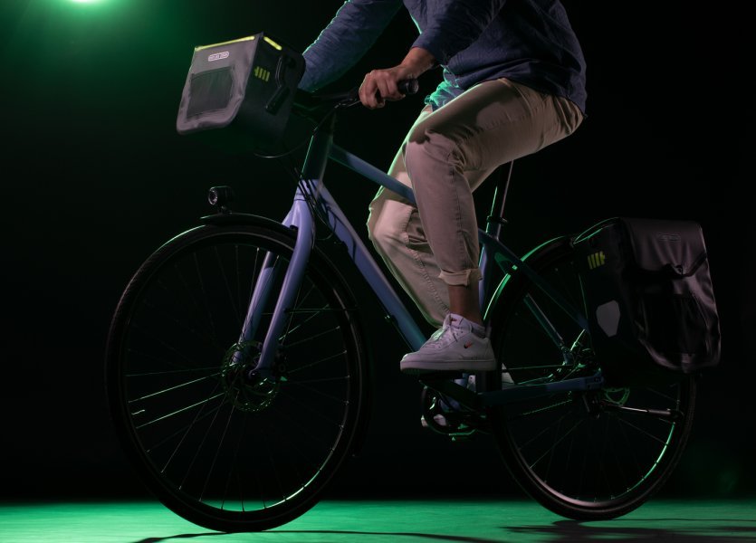 Eine Person fährt ein Fahrrad mit einer schwarzen ORTLIEB Lenkertasche mit grünen Akzenten. Die Tasche hat ein beleuchtetes Design. Eine Gepäcktasche ist hinten montiert. Der Hintergrund ist dunkelgrün.