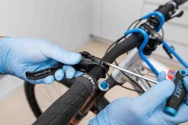 Close-Up Foto: Hände in blauen Einweghandschuhen stellen die Gebereinheit einer hydraulischen Fahrradbremse am Lenker waagerecht.