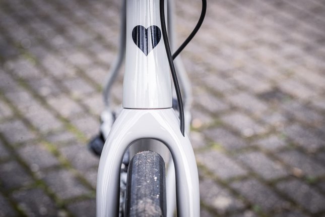 Detailfoto: Ein breiter Straßenreifen benötigt viel Platz in der Gabel eines hellgrauen Blackheart Rennrads. Das Steuerrohr ziert ein schwarzes Herz, im Hintergrund eine grau plattierte Fläche
