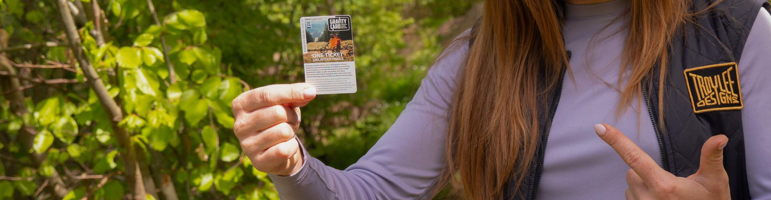 Person hält eine Gravity Card 2026 im Wald und zeigt auf das Ticket für Bikepark- und Trail-Zugang, Gewinnspielmotiv im Outdoor-Setting.