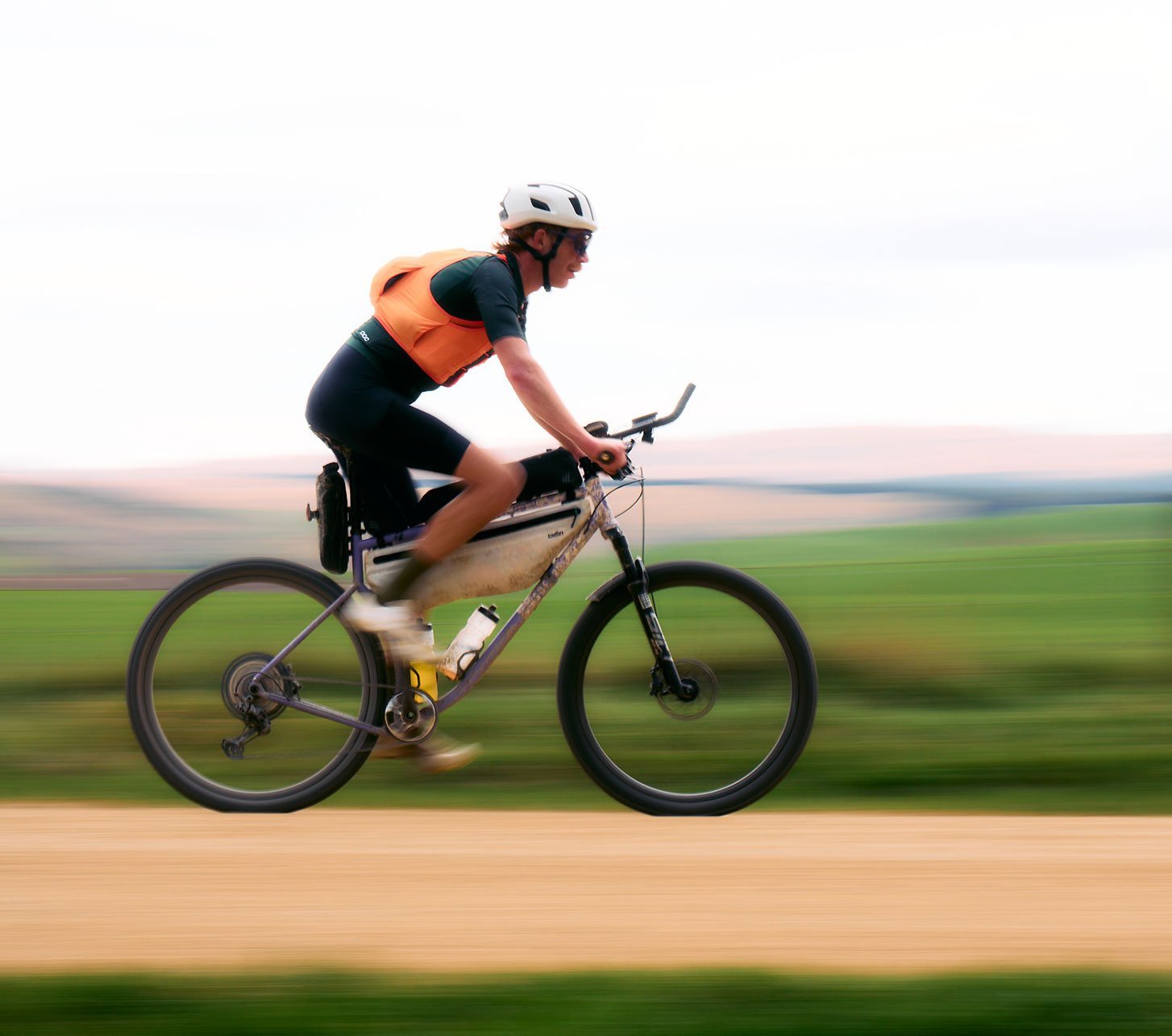 Ciclista deportivo con casco y ropa gravel POC naranja pedaleando rápido en una bicicleta gravel por un camino de tierra, fondo desenfocado que transmite velocidad.