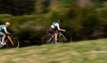 Herbert y Katrin avanzan a toda velocidad en sus bicicletas gravel por un paisaje natural verde. Toma en movimiento desde un lado, con los ciclistas enfocados y el fondo desenfocado por el movimiento.