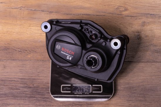 A Bosch SX e-bike drive on a kitchen scale on a dark wooden floor. Display shows 2,075 grams