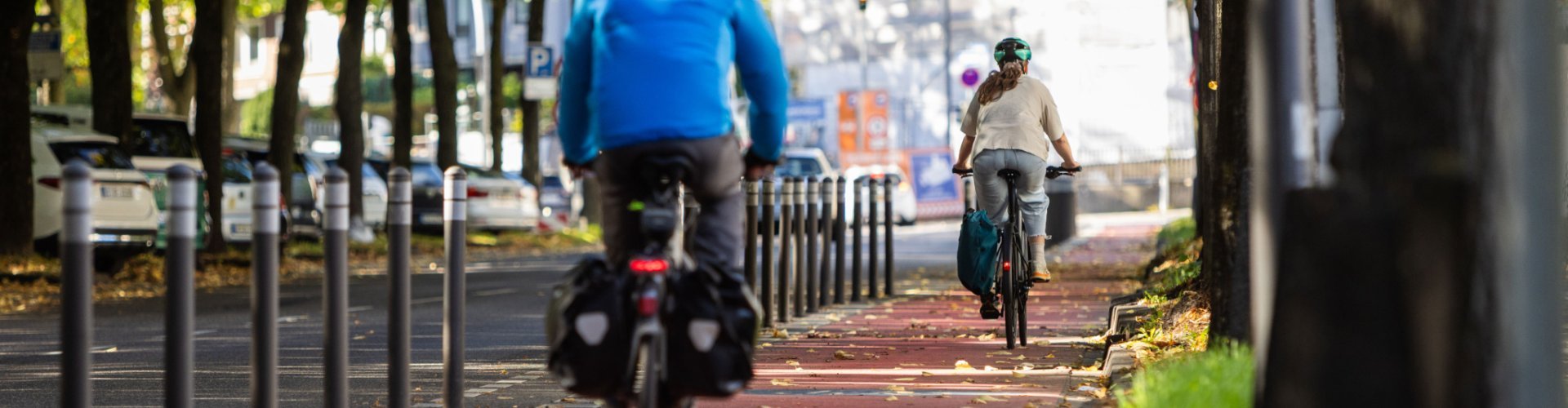 Deux cyclistes roulent sur une piste cyclable rouge bordée d’arbres ; des bornes séparent la piste de la chaussée.