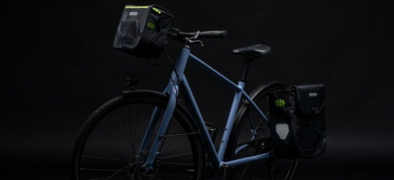Ein blaues Fahrrad mit einer schwarzen ORTLIEB Lenkertasche und einer Gepäcktasche hinten, beide mit grünen Akzenten. Die Lenkertasche hat ein beleuchtetes Design. Der Hintergrund ist dunkel.