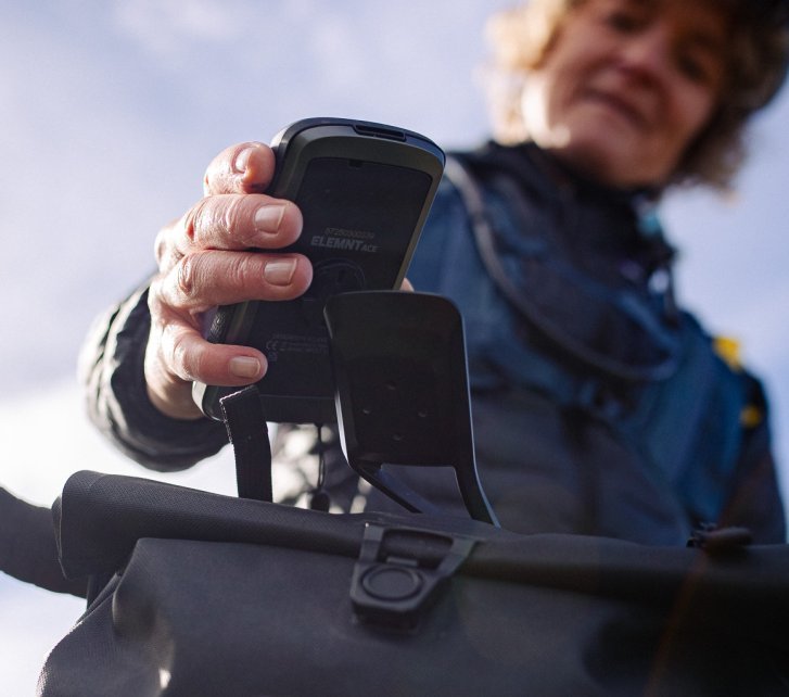 Close up von unten: Eine Frau nimmt einen Wahoo Fahrradcomputer von seiner Halterung. Wir sehen den Lenker eines Gravelbikes. Unscharf im Hintergrund das Gesicht der Frau.
