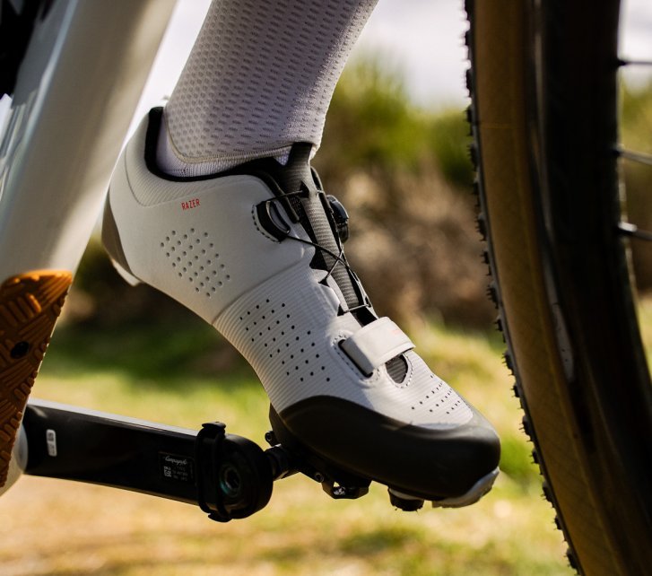 Detailfoto: Ein Fuß mit einem hellgrauen Fahrradschuh auf einem Pedal, unscharf im Vordergrund sehen wir das Unterrohr eines Fahrrads und Teile des Vorderrades.