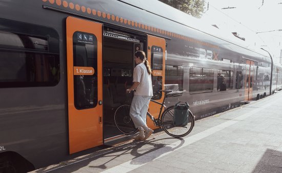 Une personne pousse un vélo équipé d’une sacoche dans un train à quai ; la porte est ouverte et d’autres voyageurs attendent en arrière-plan.