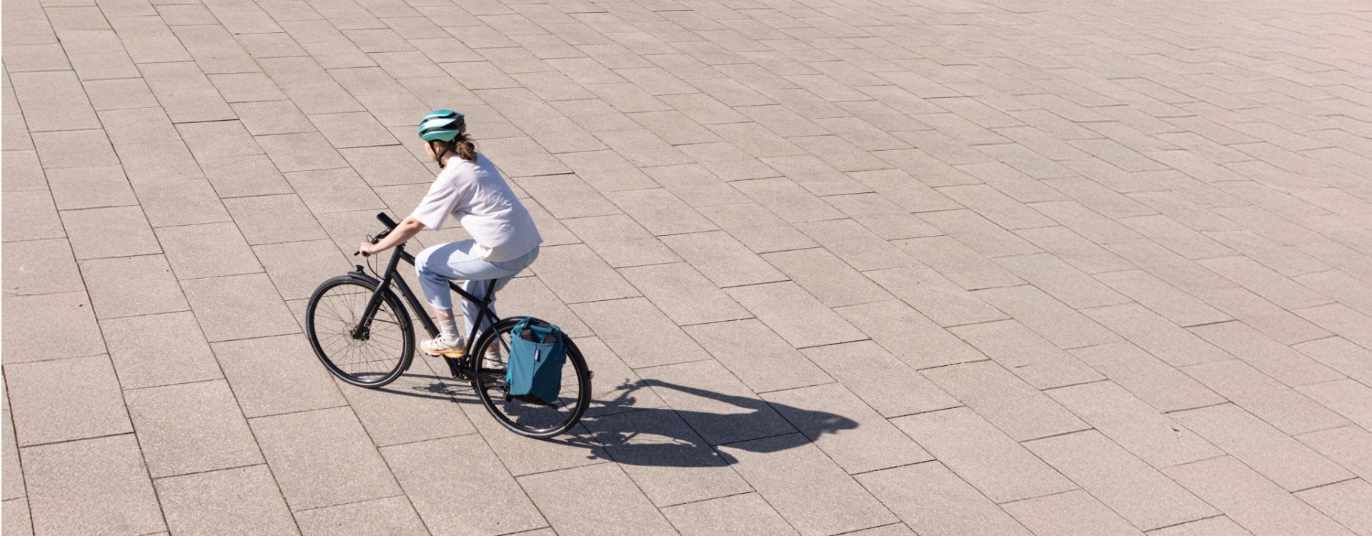 A young woman wearing a green bike helmet rides a dark city bike with a green pannier across a bright, evenly paved stone surface