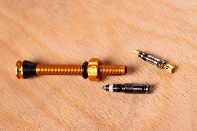 Close-up: An orange anodized tubeless valve lying on a light wooden surface, next to an SV and an SCV valve core.