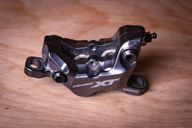 Una pinza de freno Shimano XT de 4 pistones descansa sobre una superficie de madera de haya. En su interior se distinguen claramente los pistones cerámicos de color claro.