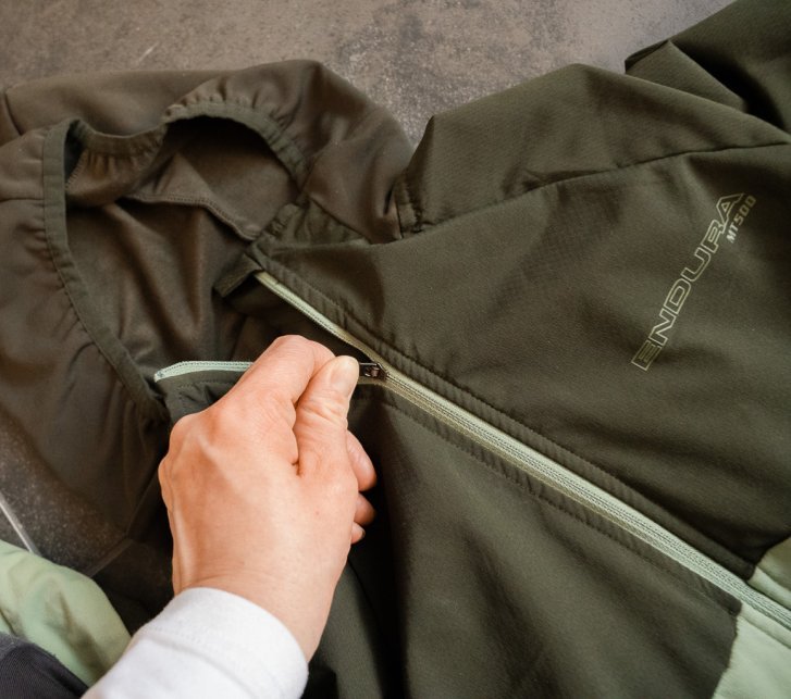 Eine grüne Funktionsjacke liegt auf einem grau gefliesten Fußboden. Eine Hand zieht den Frontreißverschluss zu.