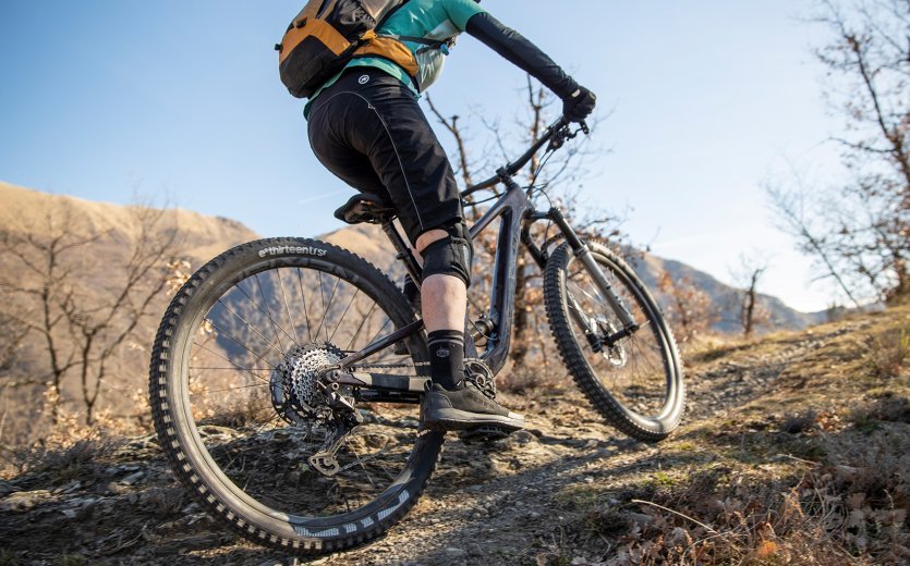 Eine Mountainbikerin fährt auf steinigem Gelände bergauf. Sie trägt einen Rucksack und Bike Wear passend zu dem frühlingshaften Wetter.