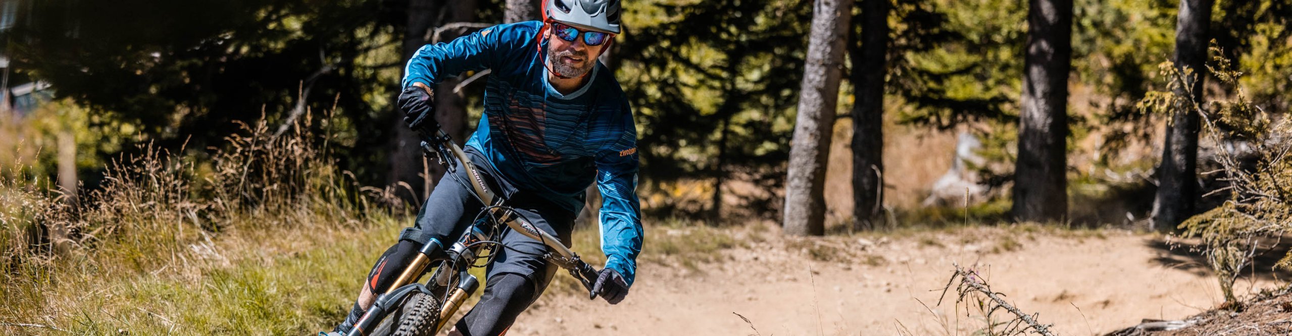 Mountainbiker in blauer Zimtstern MTB-Bekleidung fährt mit Helm und Brille eine staubige Kurve auf Waldtrail.