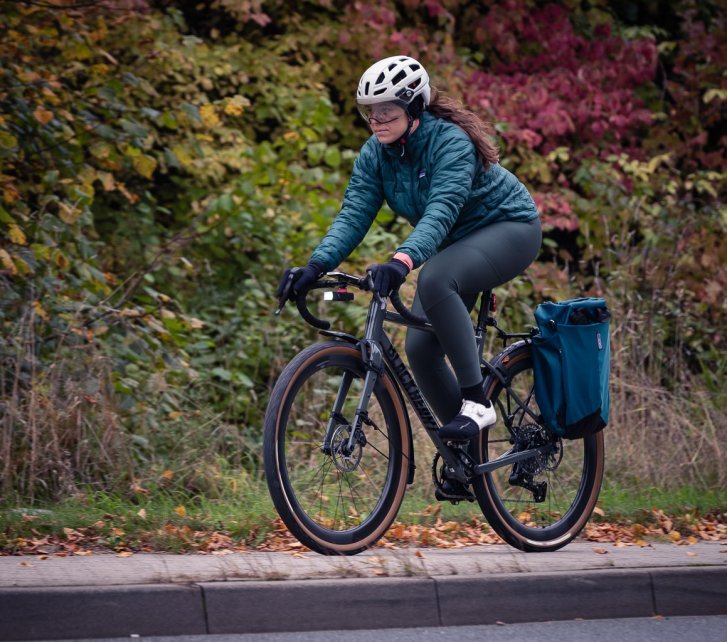 A young woman in sea-green winter clothing rides a graphite-grey gravel bike through an autumnal landscape.