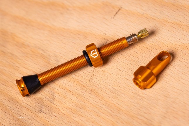 An orange tubeless bicycle valve and an orange valve cap lie on a light wooden surface