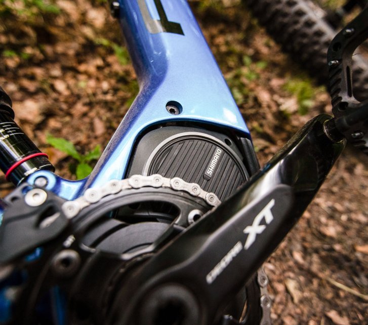 Detailfoto eines liegenden E-MTB mit blauem Rahmen. Man sieht die rechte Seite eines Shimano-Mittelantriebs und Teile von Kurbel und Kettenblatt. im Hintergrund unscharf Waldboden.