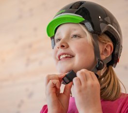 A blonde girl is fiddling with the chin strap of a black bike helmet.