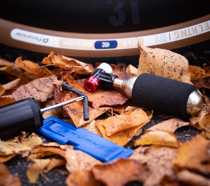 Eine CO2-Minipumpe, ein Reifenheber und ein Tubeless-Tool mit eingeklemmter Reifefensalami liegen vor einem Gravel-Reifen in buntem Herbstlaub