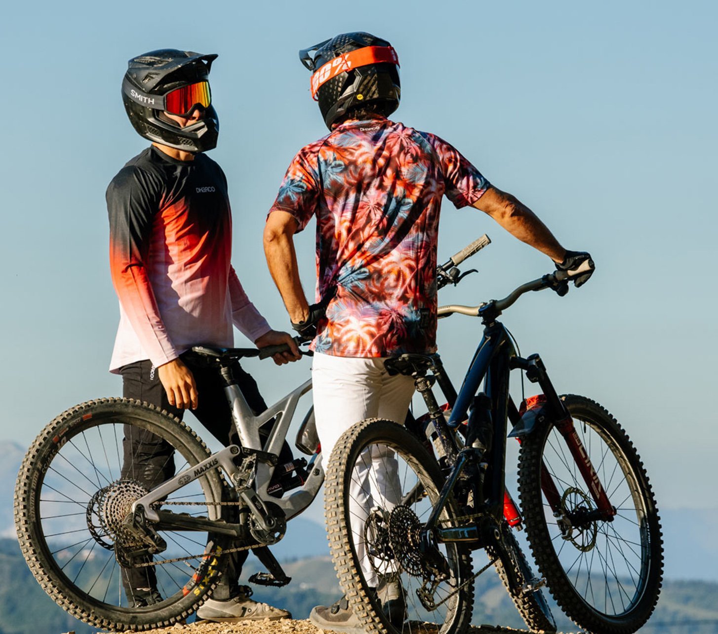 Zwei Mountainbike Fahrer stehen mit ihren MTB's vor einem wolkenlosen Alpenpanorama Hintergrund und tragen auffällige und stylische Fahrradbekleidung von DHaRCO