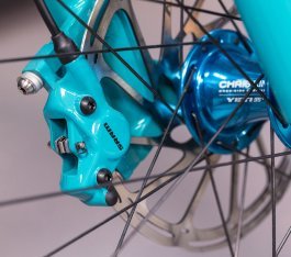 Una pinza de freno SRAM Level en un llamativo color turquesa, fotografiada a través de los radios de la rueda delantera. Se distinguen los contornos del único pistón interior dentro de la pinza.