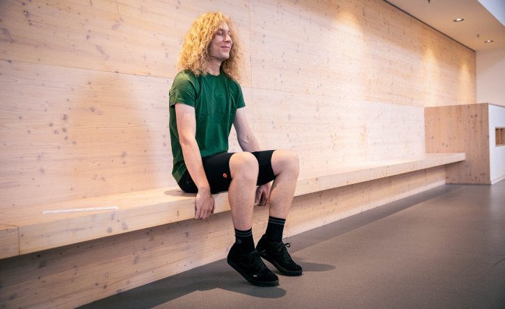 A person sits grinning on a modern, light wooden bench on a piece of corrugated cardboard, pulling themselves firmly onto the seat with their hands.
