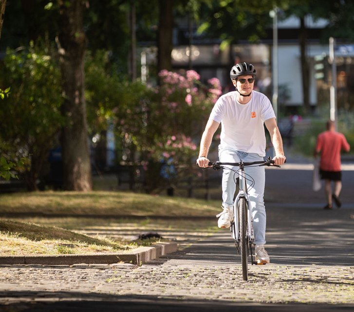 A person rides a bicycle on a paved bike path in an urban setting, surrounded by trees and a relaxed atmosphere, while another person goes in the background.