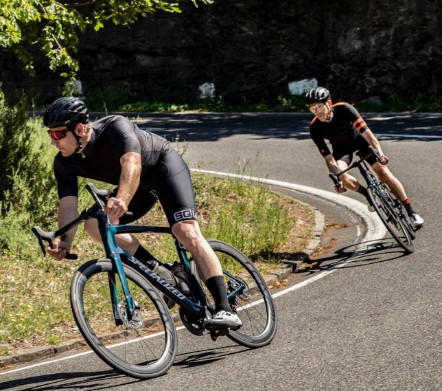 Two cyclists in black sportswear with helmets are riding on a winding road through a green landscape. The leading cyclist is on a Specialized bike, while the other follows behind. The road is surrounded by grass and trees.