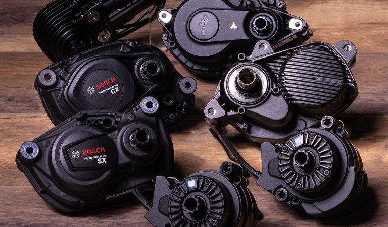 Sieben verschiedene, schwarze E-Bike Antriebe liegen auf einer dunklen Holzoberfläche