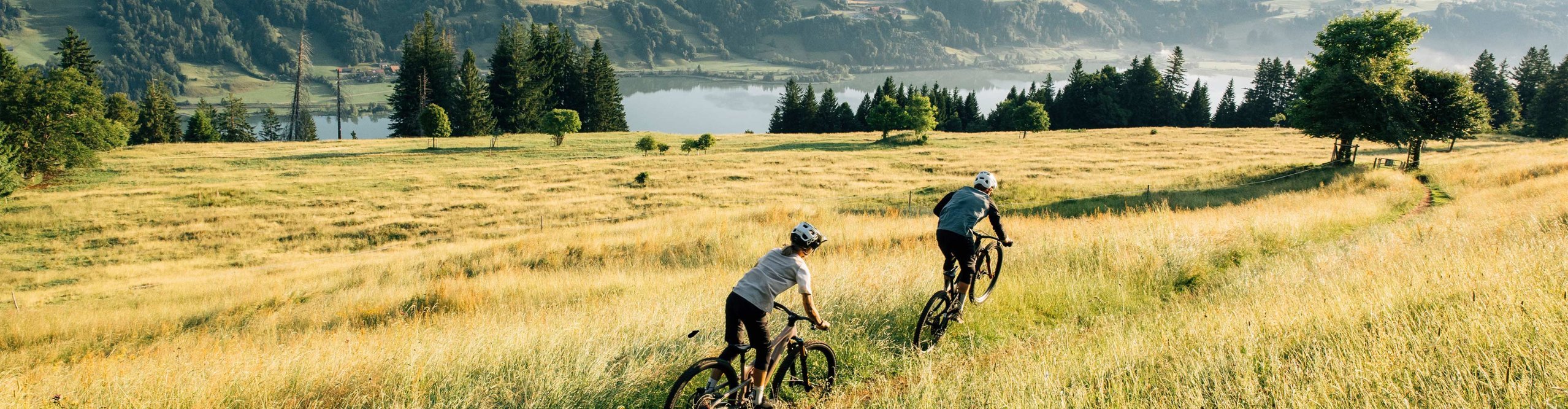 Zwei Mountainbiker in VAUDE Fahrrabekleidung fahren mit ihren Mountainbikes über eine hohe Feldwiese - im Hintergrund Alpines Talgelände und ein Bergsee.
