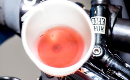 Ein weißer Trichter, gefüllt mit rotem Mineralöl, ist an einen Bremshebel eines Fahrrads angeschlossen, mit einer "RockShox"-Aufschrift am Lenker sichtbar.