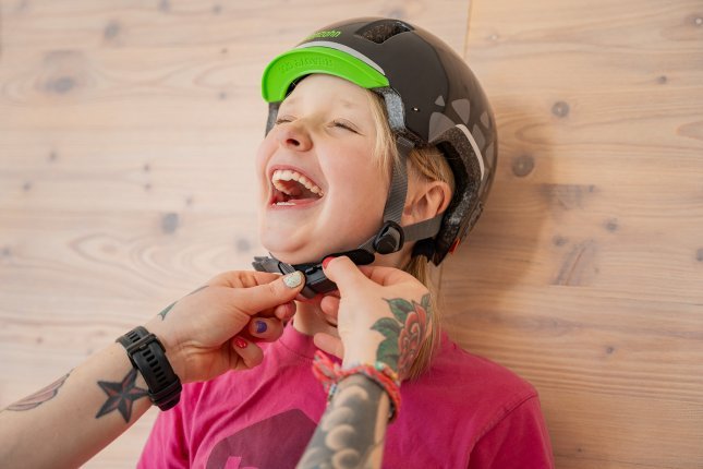 The colorful tattooed hands of an adult woman are fastening the chin strap of a bike helmet around the neck of a blonde girl.