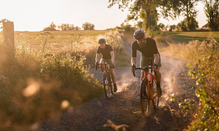 Deux cyclistes gravel roulent sur un chemin poussiéreux au coucher du soleil, poussière visible en contre-jour.