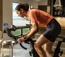 Una mujer de pelo oscuro entrena en una bici indoor en una cocina-salón de tonos cálidos y aspecto elegante.