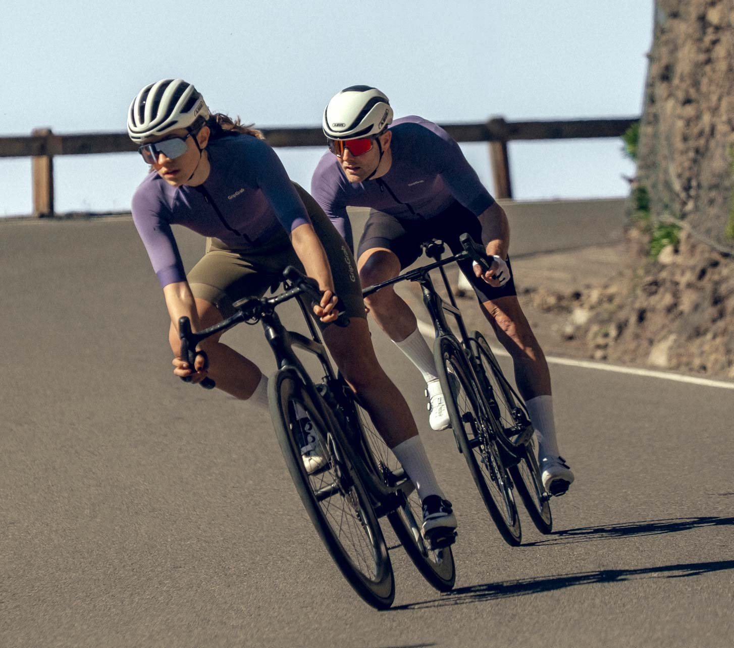 Dos ciclistas de carretera con ropa de ciclismo GripGrab toman una curva rápida en una carretera de montaña soleada.