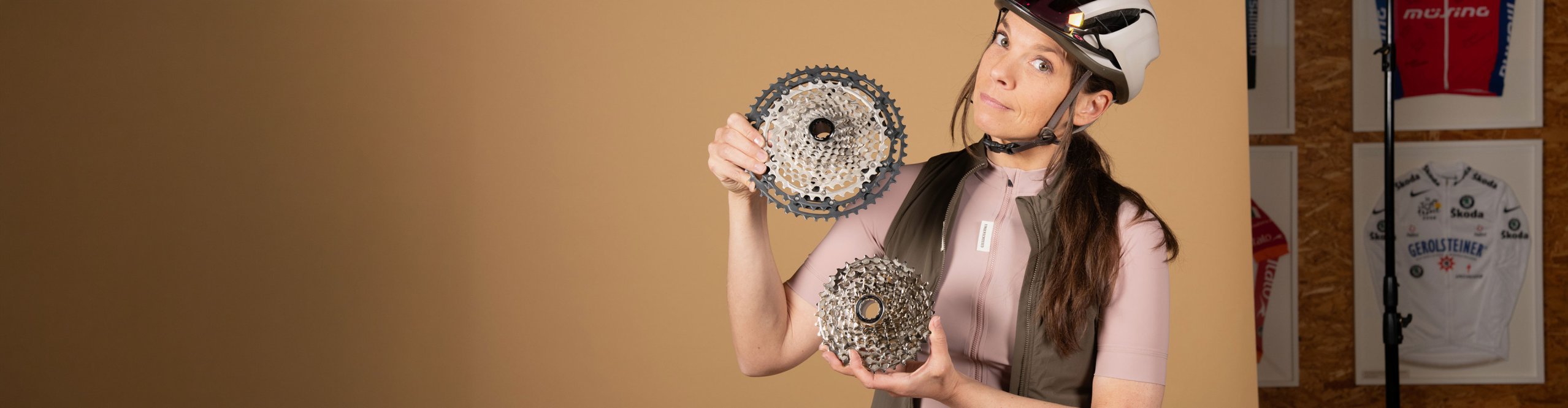 Radfahrerin Anke mit Helm hält zwei unterschiedlich große Fahrradkassetten in den Händen und vergleicht sie vor neutralem Hintergrund