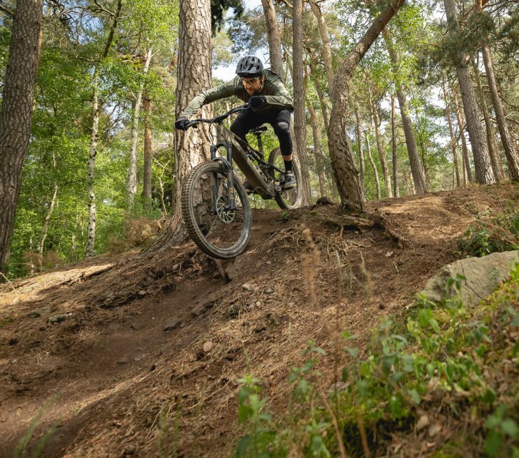 Berno heizt mit einem bronze-grünen E-MTB einen Trail im Wald hinunter 