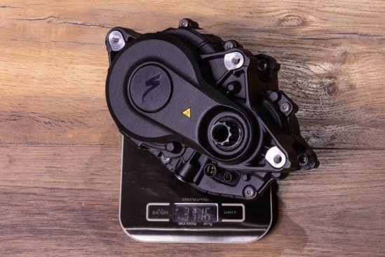 A Specialized 3.1 e-bike drive on a kitchen scale on a dark wooden floor. Display shows 3,116 grams
